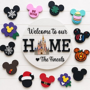May include: A round white wooden sign with the text Welcome to our HOME featuring a castle illustration and the name The Fincels. It is surrounded by twelve interchangeable Mickey Mouse-shaped icons representing various holidays and seasons.