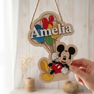 May include: Wooden sign featuring Mickey Mouse holding balloons, with the name "Amelia" in white letters. The sign has a natural wood finish and is suspended by a rope. The balloons are red, blue, yellow, and green.