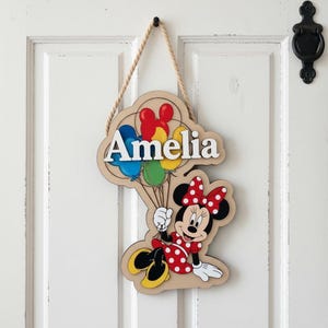 May include: Wooden door sign featuring the name "Amelia" in white, with a cartoon Minnie Mouse holding colorful balloons. The sign is suspended by a rope and has a Minnie Mouse design with a red polka dot dress and bow.