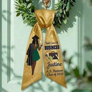Op de afbeelding: Gouden afstudeerlint met de tekst "Took Care Of Business" en "Justine A.S. Business Class of 2026". Het lint toont een illustratie van een afgestudeerde in toga en baret. Bovenop een groene krans.