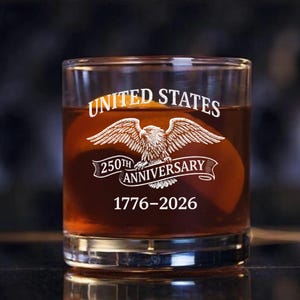 Może przedstawiać: Przezroczysty szklany tumbler wypełniony brązowym płynem. Na szkle wygrawerowano napisy "UNITED STATES" nad emblematem orła oraz "250TH ANNIVERSARY 1776-2026" poniżej. Szklanka jest prawdopodobnie przeznaczona do whisky lub innych alkoholi.