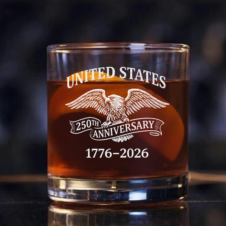 Może przedstawiać: Przezroczysty szklany tumbler wypełniony brązowym płynem. Na szkle wygrawerowano napisy "UNITED STATES" nad emblematem orła oraz "250TH ANNIVERSARY 1776-2026" poniżej. Szklanka jest prawdopodobnie przeznaczona do whisky lub innych alkoholi.