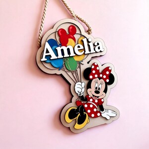 May include: A wooden sign with the name "Amelia" in white, and a cartoon Minnie Mouse holding balloons. The sign has a beige cloud-shaped background and a rope for hanging. Minnie Mouse is wearing a red polka-dot dress.