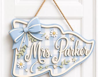 May include: A decorative wall sign featuring a light blue and white gingham pattern with the text Mrs. Parker in white script. The sign is adorned with a large light blue bow, small white daisy flowers, stars, and green leaf accents, suspended by a brown rope.