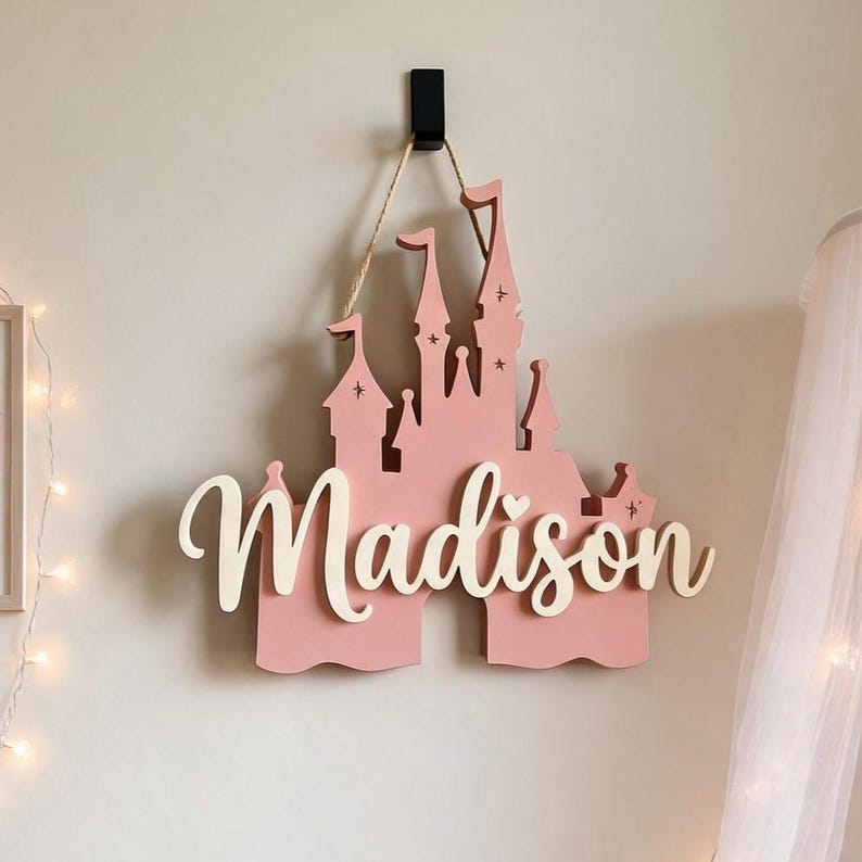 Pode incluir: Uma placa de madeira rosa em forma de castelo com o nome "Madison" em escrita branca. A placa est&aacute; pendurada em uma parede por um gancho preto e barbante. O castelo tem v&aacute;rias torres e elementos decorativos.