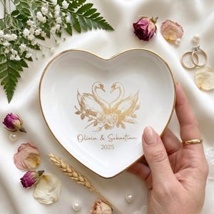 Pode incluir: Uma tigela de anel de cerâmica branca em forma de coração com uma borda dourada. A tigela apresenta uma ilustração dourada de dois cisnes e detalhes florais. Os nomes "Olivia & Sebastian" e o ano "2025" são impressos.