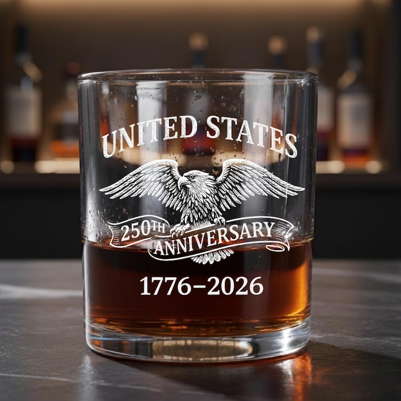 Może przedstawiać: Przezroczysta szklanka do whisky wypełniona bursztynowym płynem. Na szklance widnieje napis "UNITED STATES" nad wygrawerowanym orłem. Poniżej znajduje się napis "250TH ANNIVERSARY" oraz daty "1776-2026".