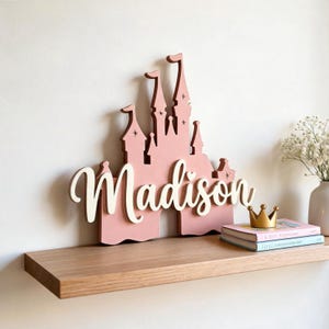 Pode incluir: Uma placa de madeira rosa em forma de castelo com o nome "Madison" em escrita branca. A placa &eacute; exibida em uma prateleira de madeira, ao lado de uma coroa de ouro e livros empilhados. O castelo tem v&aacute;rias torres e detalhes de estrelas.
