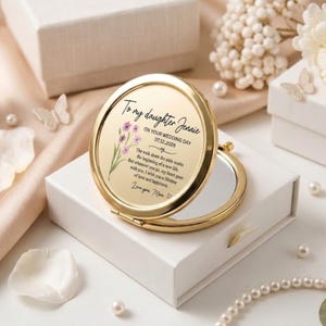 Pode incluir: Um espelho compacto dourado com um design floral e o texto "To my daughter Jennie ON YOUR WEDDING DAY 07.12.2026". O espelho está aberto, revelando uma superfície refletora. O compacto está numa caixa branca.