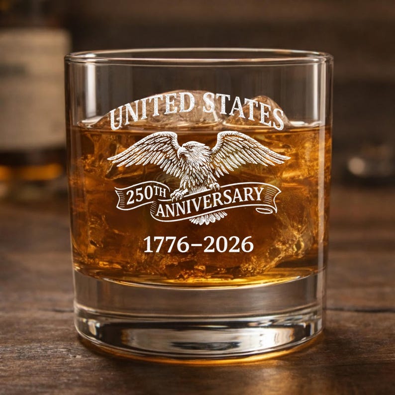 Może przedstawiać: Przezroczysta szklanka do whisky wypełniona bursztynowym płynem i kostkami lodu. Na szklance wygrawerowano napisy "UNITED STATES" nad motywem orła oraz "250TH ANNIVERSARY 1776-2026". Szklanka stoi na drewnianej powierzchni.