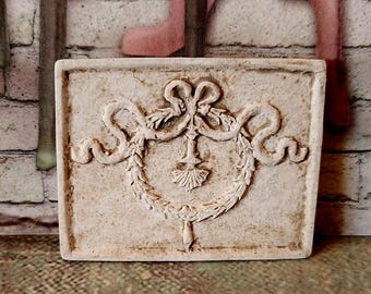 May include: A rectangular, off-white, decorative plaque with a raised relief design. The design features a central wreath, a bow, and flowing ribbons. The plaque has a textured surface and a slightly aged appearance, suggesting an antique or vintage style.
