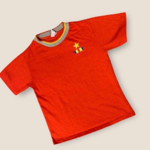 Könnte beinhalten: Ein leuchtend rotes T-Shirt mit grauem und gelbem Kragen. Ein kleines gesticktes Emblem mit einem goldenen Stern und bunten Streifen befindet sich auf der linken Brust. Das Shirt hat kurze Ärmel und ein leicht strukturiertes Aussehen.