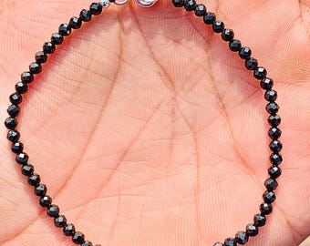 May include: A delicate black beaded bracelet with a silver clasp. The bracelet is composed of small, faceted black beads strung together. The clasp is a simple silver design, and the bracelet is displayed on a person's hand.