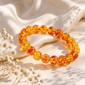 Peut inclure: Un bracelet composé de perles d'ambre. Les perles rondes sont d'une couleur jaune doré translucide avec des inclusions rouges et oranges. Le bracelet est posé sur un tissu blanc et soyeux.