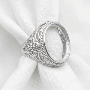 May include: A silver-coloured metal ring setting featuring an oval-shaped centre bezel surrounded by a beaded border. The band and sides of the ring are decorated with intricate, raised floral and scrollwork patterns in a metallic finish.