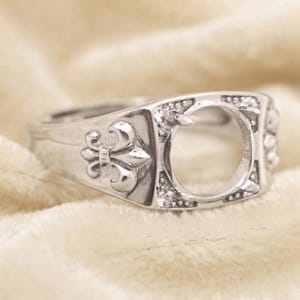 Könnte beinhalten: Ein silberfarbener Metallring mit einer zentralen kreisförmigen Fassung für einen Stein, flankiert von Fleur-de-Lis-Motiven an den Seiten. Der Ring ist mit kleinen, klaren, funkelnden Steinen besetzt, die um die kreisförmige Fassung herum liegen.