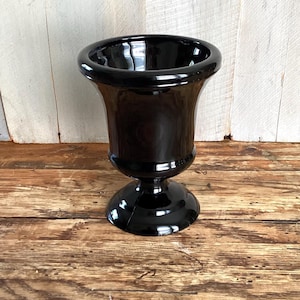 May include: A black glass urn with a flared rim and a pedestal base. The urn is made of glossy black glass and sits on a wooden surface. The urn is a decorative item.