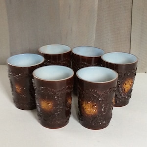 May include: Set of six brown and white ceramic drinking glasses with a raised floral design. The glasses have a light brown glaze with a darker brown floral pattern.