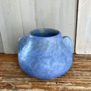 May include: A blue ceramic vase with two handles. The vase has a rounded body and a textured surface. The color is a muted blue with hints of green and gray. The vase is sitting on a wooden surface.