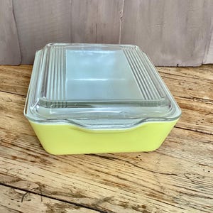 May include: A vintage yellow rectangular Pyrex baking dish with a clear glass lid. The lid has vertical ribbed detailing. The dish is sitting on a wooden surface.