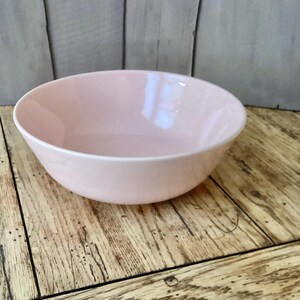 May include: A light pink ceramic bowl with a white rim. The bowl is set on a wooden surface with a light gray wooden background. The bowl is empty and appears to be new.