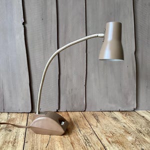 May include: A vintage desk lamp with a beige shade and base, connected by a flexible, silver-colored gooseneck arm. The lamp sits on a wooden surface, and the cord extends to the left. The lamp's design is reminiscent of mid-century modern aesthetics.