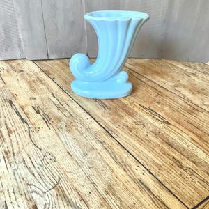 May include: A light blue ceramic vase with a horn-like shape and a scalloped rim. The vase has a curved, scroll-like design on its base. The vase is displayed on a wooden surface.