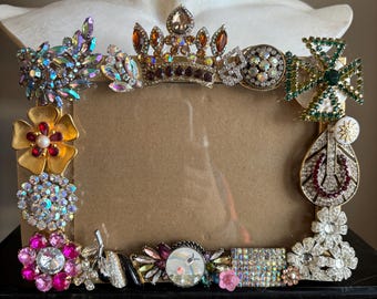 May include: Ornate picture frame embellished with a variety of vintage brooches and jewels. The frame features a crown, flowers, and other decorative elements in shades of gold, pink, green, and iridescent colors. The frame is designed to hold a rectangular photo.