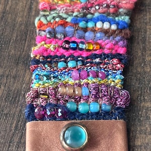Könnte beinhalten: Ein gewebtes Textilarmband mit einer lebendigen Auswahl an mehrfarbigem Garn, Metallic-Fäden und verschiedenen facettierten Glasperlen in Blau-, Rosa-, Orange- und Violetttönen. Das Armband wird mit einer hellbraunen Lederlasche und einem blauen Druckknopf verschlossen.
