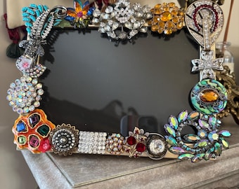 May include: A rectangular photo frame with a black interior, embellished with a variety of colorful vintage brooches. The brooches feature rhinestones, crystals, and various jewel tones, creating a unique and ornate design.
