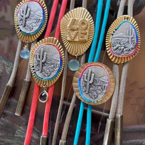 May include: A collection of five bolo ties featuring silver-toned oval or circular medallions with embossed desert cactus landscapes or the word LOVE in gold-toned metal. Each medallion is framed by colorful beads and suspended on braided cords.
