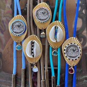May include: Five bolo ties featuring gold-toned, sunburst-shaped metal backings. Three ties display silver-toned circular wave reliefs, while two feature white cowrie shells. Each includes a small, dangling faceted gemstone and metal tips.