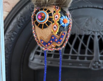 Könnte beinhalten: Handgefertigter Eulen-Ornament aus Filz mit einem Kunstfellkopf. Die Eule hat ein braunes und schwarzes Gesicht mit farbenfrohen Stickereidetails, ein großes, mit Juwelen besetztes Knopfauge und blaue, mit Perlen besetzte Beine. Die Eule hängt an einem Holzdübel mit farbenfrohen gewebten Armbändern.