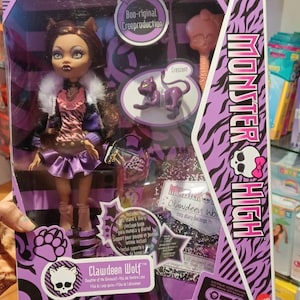 Può includere: Una bambola Clawdeen Wolf di Monster High in una scatola viola e nera. La bambola ha capelli castani, orecchie da gatto e un abito viola e rosa. La scatola presenta il logo Monster High e il testo "Boo-riginal Creeproduction."