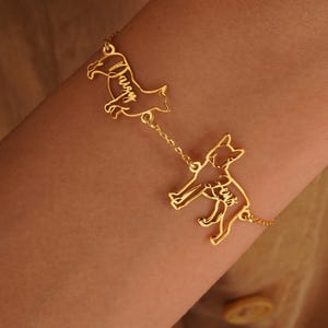 May include: Gold-colored bracelet featuring two dog silhouettes connected by a delicate chain. Each dog has a name engraved on it. The bracelet is worn on a wrist, showcasing its design and craftsmanship.