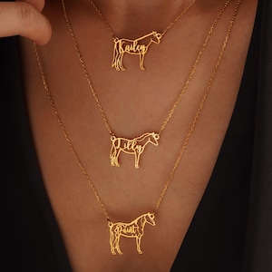 May include: Three gold-colored necklaces with horse pendants. Each pendant features a horse silhouette and a name: "Bailey", "Dilly", and "Paint". The necklaces are layered, creating a tiered effect. The background is a dark, solid color.