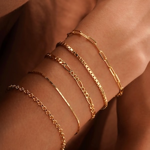 May include: A collection of gold bracelets in various chain styles. The bracelets include a delicate chain, a box chain, a Figaro chain, a curb chain, and a paperclip chain. The jewelry is displayed on a person's arm.