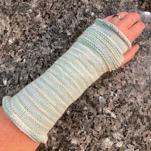 Może przedstawiać: Dzianinowy ocieplacz na ramię w odcieniach jasnoniebieskiego i białego. Ocieplacz na ramię ma prążkowany mankiet i wzór w paski na ramieniu. Artykuł jest ręcznie robiony i wydaje się miękki i wygodny.