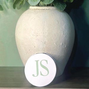 May include: A white, round compact mirror with the initials "JS" in a light green colour. The mirror is placed in front of a large, off-white vase with a textured surface. Green foliage is visible at the top of the vase.