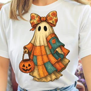 以下が含まれることがあります： 白いTシャツに漫画の幽霊のデザイン。幽霊はパッチワークの衣装、花の弓を着て、ジャック・オー・ランタンのバケツを持っています。デザインは秋の色で、ハロウィーンに適しています。
