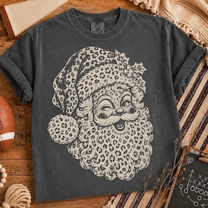 May include: A charcoal grey short-sleeve t-shirt featuring a large, cream-coloured graphic of a winking Santa Claus face. The Santa hat and beard are decorated with a detailed leopard print pattern, and a sprig of holly sits on the hat.