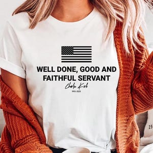 May include: White t-shirt featuring a black and white American flag graphic above the text "WELL DONE, GOOD AND FAITHFUL SERVANT" and the name "Charlie Kirk" with the years 1993-2025. The shirt has short sleeves.