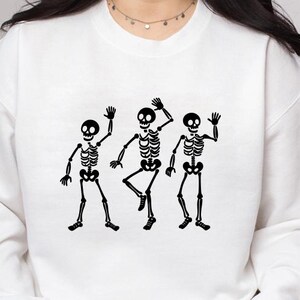 Puede incluir: Sudadera blanca con un gr&aacute;fico negro de tres esqueletos bailando. Los esqueletos est&aacute;n representados en movimiento, con los brazos levantados y las piernas en varias posiciones. El dise&ntilde;o est&aacute; centrado en la parte delantera de la sudadera.
