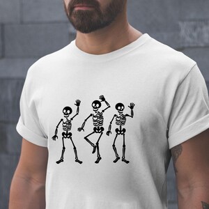 Puede incluir: Camiseta blanca con un gr&aacute;fico negro de tres esqueletos bailando. Los esqueletos est&aacute;n representados con cajas tor&aacute;cicas, extremidades y cabezas redondas detalladas, saludando y bailando. La camiseta es de material suave.