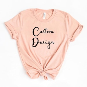 Puede incluir: Una camiseta rosa claro con mangas enrolladas y dobladillo anudado. Las palabras "Custom Design" est&aacute;n escritas en cursiva negra en la parte delantera. La camiseta est&aacute; hecha de un material suave.