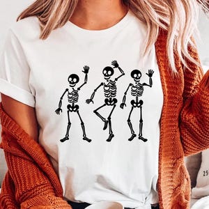 Puede incluir: Camiseta blanca con un gr&aacute;fico negro de tres esqueletos bailando. Los esqueletos est&aacute;n representados en diferentes poses, con los brazos levantados. La camiseta es de estilo casual, adecuada para Halloween o eventos tem&aacute;ticos.