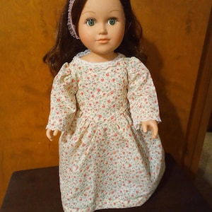 May include: A doll with long brown hair, green eyes, and a light pink headband with lace trim. The doll wears a long-sleeved, floor-length dress featuring a small floral pattern of tiny red flowers and green leaves on a cream-colored background.