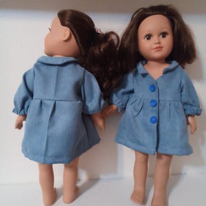 May include: Two dolls with brown hair wearing matching blue denim-style coats with three blue buttons. One doll is facing forward, the other is turned to the side. The dolls are standing on a white surface.