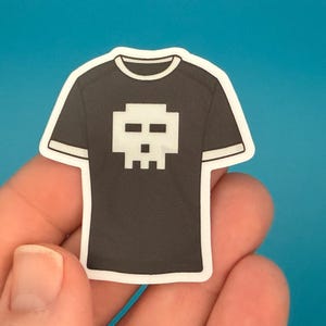 May include: A black t-shirt sticker with a white skull design. The sticker has a white border and is held in a person's hand. The t-shirt design is a pixelated skull.