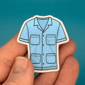 May include: A light blue sticker featuring a short-sleeved shirt design. The shirt has a collar, two chest pockets, and two lower pockets. The sticker is held in a person's hand against a blue background.